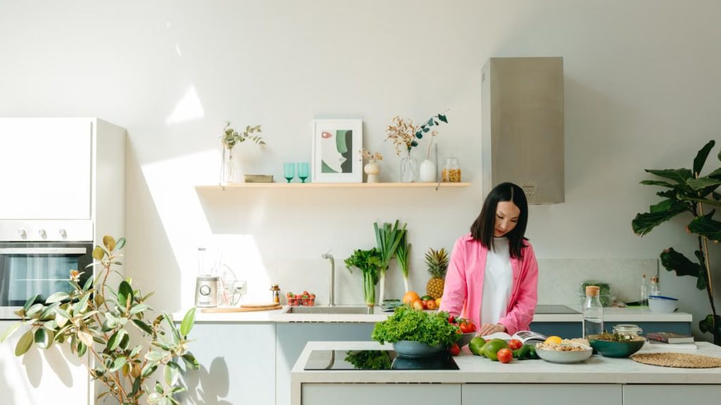 A woman in pink prepares fresh vegetables in a sunlit, modern kitchen, creating a vibrant cooking scene.